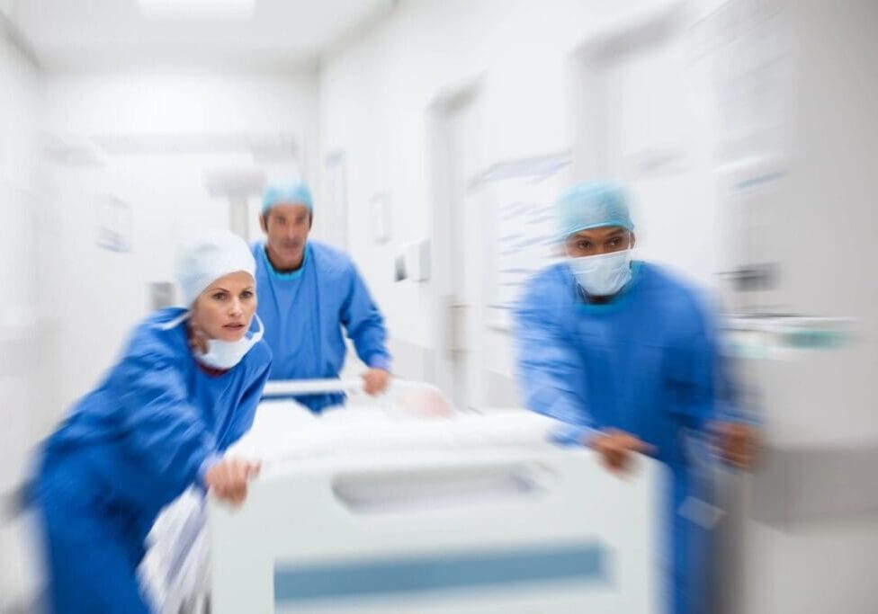 Three doctors in blue gowns and masks are pushing a bed.